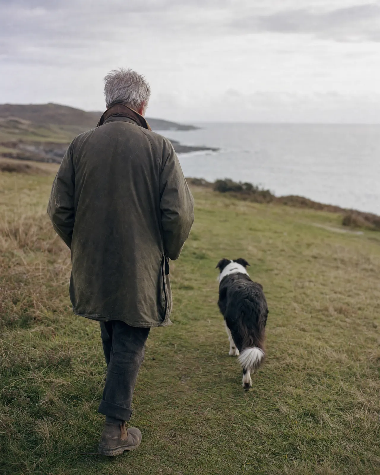 An older man walking slowly with a black-and-white collie along a coastal path